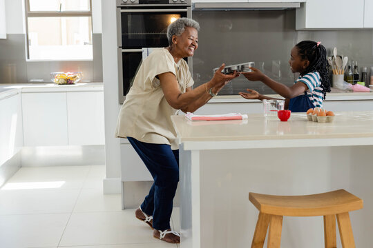 Senior African grandmother passing round pan toward child granddaughter at modern kitchen island