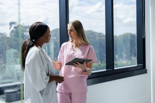 Female nurse in scrubs touching African American patient in gown showing tablet by clinic window