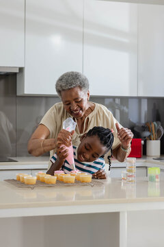 African grandmother guiding granddaughter, squeezing pink icing bag at kitchen island with cupcakes