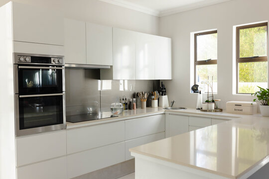Modern white kitchen with glossy island and built-in ovens is glowing in daylight, showing utensils