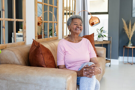 Senior African woman sitting on tan leather sofa in living room, smiling with wrist device
