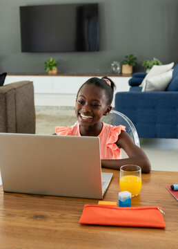 Girl child wearing coral ruffled top sitting at wooden table in living room using silver laptop
