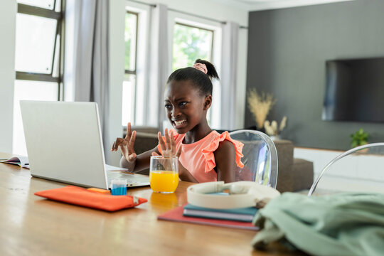 Adolescent African girl in pink ruffle top gesturing frustrated at table at home with silver laptop