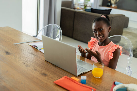 African American child gesturing toward silver laptop at home dining table in coral top, copy space