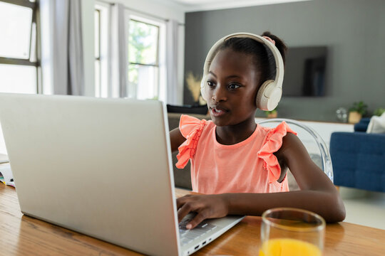 African American child sitting at home table wearing coral top using laptop with headset and juice