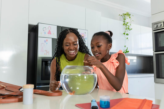 African mom, youth daughter in green tank, coral top feeding goldfish in fishbowl on kitchen island