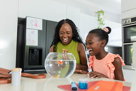 African American mom and child laughing and leaning over fishbowl with orange goldfish in kitchen
