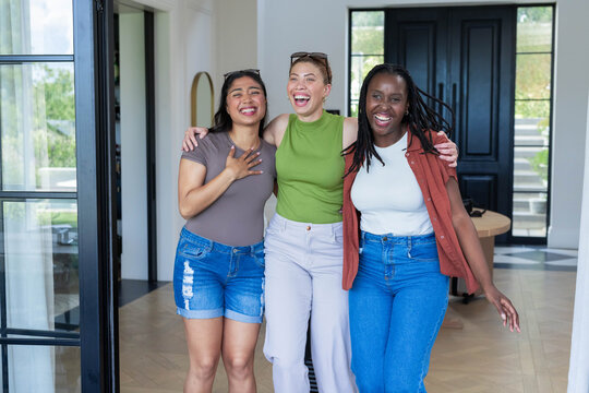 Diverse female friends laughing and hugging in entryway, wearing sunglasses near glass sliding door