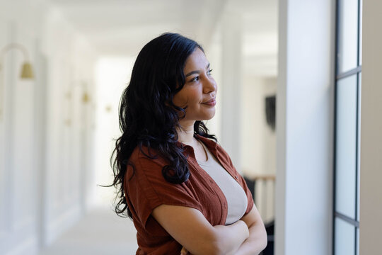 Adult female standing near tall window in bright corridor, looking contemplative with arms crossed