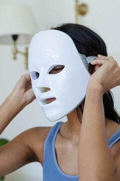 Glossy white LED facial mask is being adjusted with elastic strap near brass wall lamp