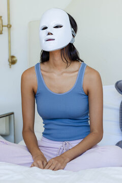 Woman is sitting cross-legged on bed wearing white face mask, blue tank top, lilac pants