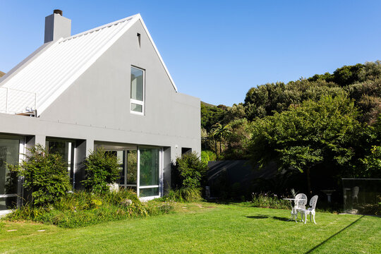 Modern two-story gray house is sitting left of frame on manicured lawn with white bistro set
