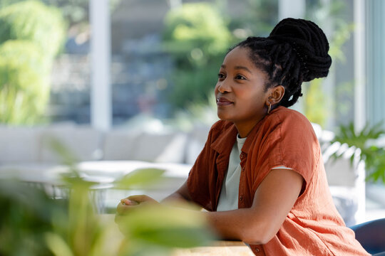 African woman in rust shirt and earrings, sitting at wooden table in bright lounge with plants