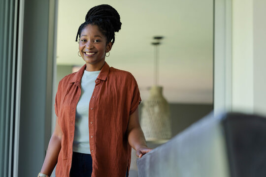 Adult African woman standing by tall window, resting on sofa arm, wearing rust shirt and wristwatch