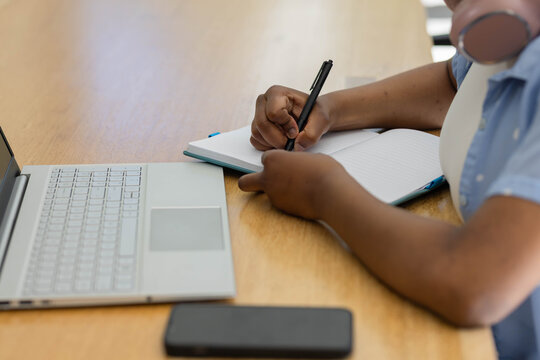African female youth writing in lined notebook at table, using black pen, silver laptop, headphones
