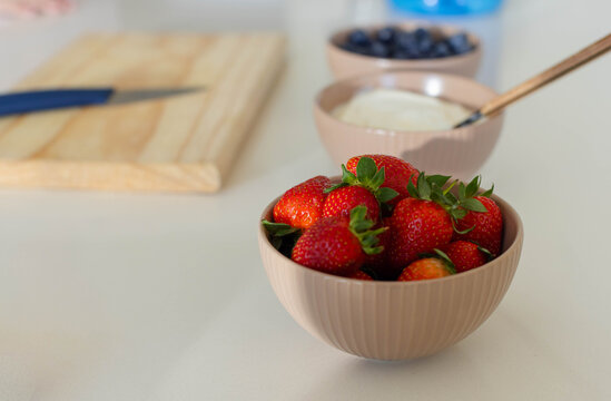 Beige-pink bowl is holding ripe strawberries on countertop with blueberries and spoon blurred