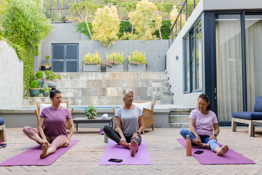 Diverse female friends in sportswear doing seated yoga on patio with purple mats, phones