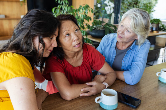 Mature women friends leaning in, comforting red-blouse friend at cafe with turquoise mugs and phone