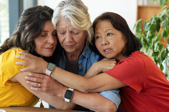Diverse female friends hugging and comforting senior woman in denim shirt at home, showing watches