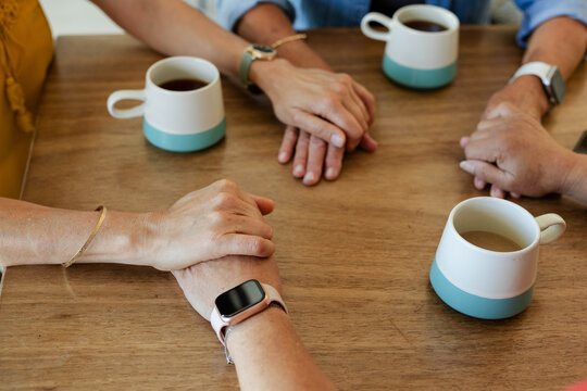 Interlocked adult hands are resting on wooden tabletop near three teal-based coffee mugs