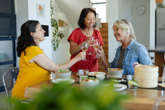 Clinking wine glasses, diverse female friends sitting around wooden dining table in home kitchen