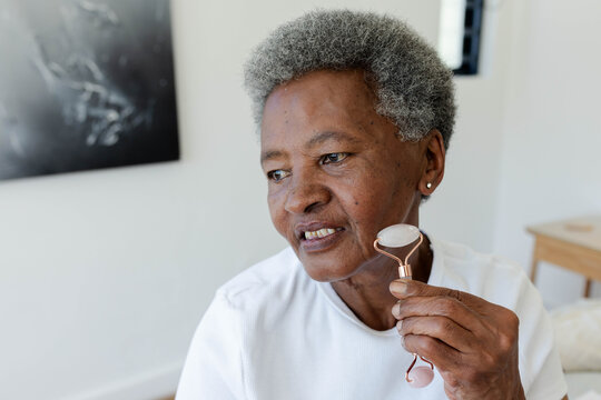 Senior African woman wearing white top using pink facial roller, rolling jawline near bedside table