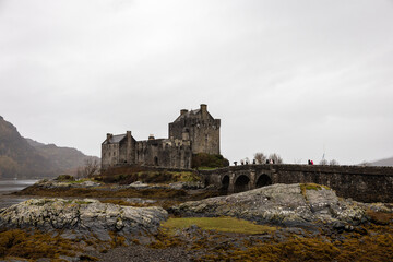 Fototapeta premium Historic stone castle standing on a rocky landscape under an overcast sky, featuring traditional medieval architecture and a stone bridge leading to the entrance. The scene evokes a dramatic and mood