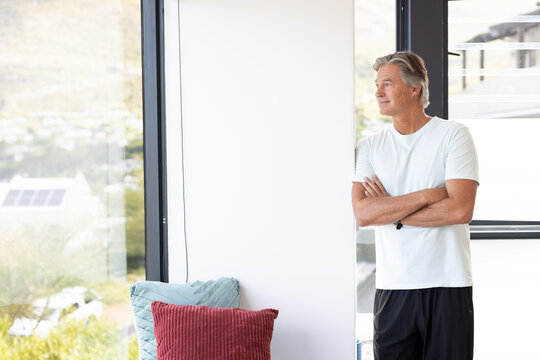 Senior man standing in living-room at right window, wearing light tee and dark shorts, copy space