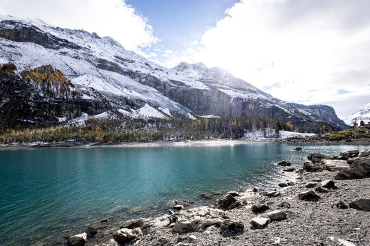 lago alpino de &aacute;guas turquesa (cor glacial t&iacute;pica, causada por part&iacute;culas minerais finas suspensas na &aacute;gua), com margem pedregosa/rochosa, cercado por montanhas nevadas ou com neve parcial nos picos e