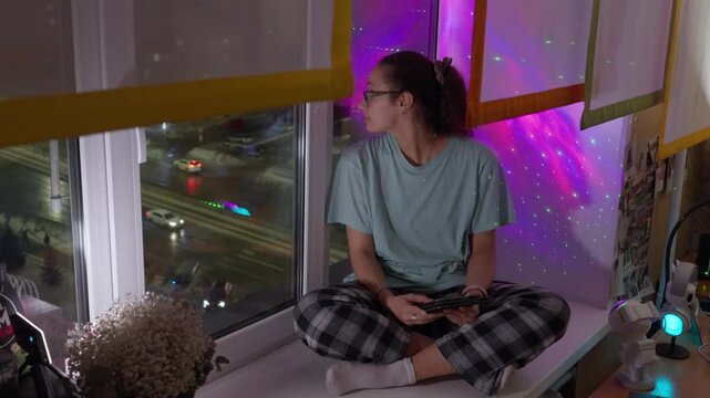 Caucasian Woman Reading At Window, Gazing At City Lights With Nebula Projection Behind Crosslegged On Sill In Plaid Pants, Hands Folded On Tablet, Thoughtful Calm During Late Evening