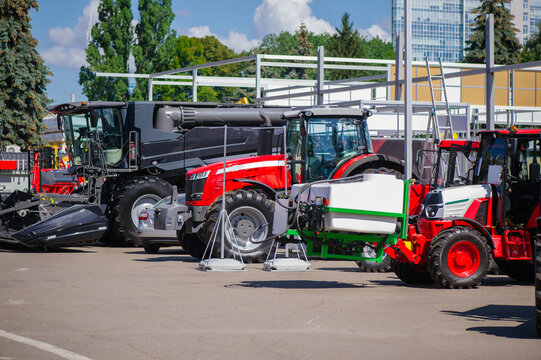 A wide-angle shot of an outdoor exhibition featuring powerful farming equipment. In the frame are a large black combine harvester and several red and white modern tractors parked on an asphalt lot und