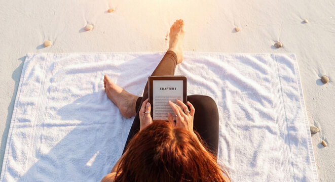 Young redheaded woman sitting on white towel at sandy beach reading digital book on electronic reader during summer vacation while enjoying bright sun and peaceful relaxation in tropical paradise.