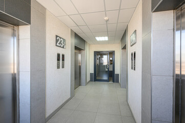 Modern elevator hallway with light grey tiled floor, textured walls, stainless steel elevators, numbered signs (23), glass door at end, recessed ceiling lights, clean minimalist design © evgeniykleymenov