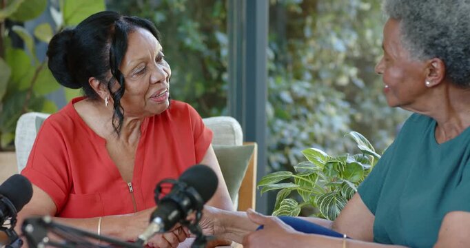 Two Senior women offering fist bump, holding hands and podcasting into microphones in living room