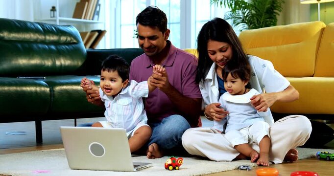 Indian Asian young parents dancing with twin infant babies to music on laptop screen during morning routine at home, engaging kids in fun audio visual learning activity with joyful family bonding