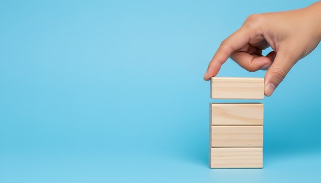 A hand carefully places a single wooden block on top of a stack of four other wooden blocks