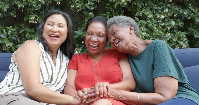 Diverse female friends seniors center red zip dress laughing after joke holding hands on blue sofa