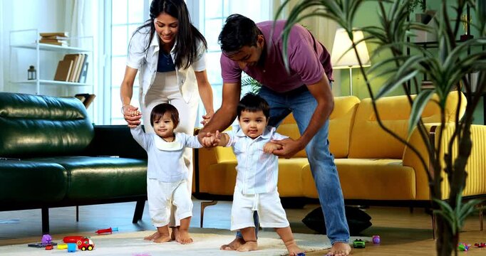 Indian Asian young parents helping twin infant babies walk by holding their hands inside modern home living room, guiding first steps together as smiling babies enjoy playful family bonding moment