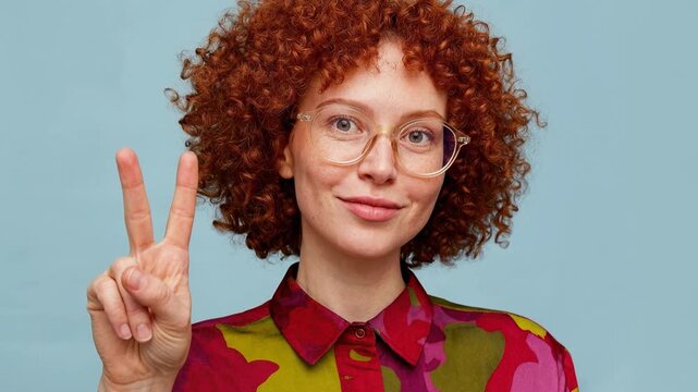 Studio portrait of cheerful red haired woman with freckles showing peace gesture against soft background highlighting individuality positivity and vibrant modern style