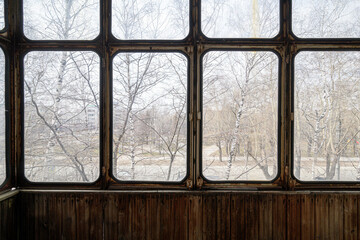Fototapeta premium A weathered multi-pane window frames a bleak winter scene: bare birch trees, a foggy street, distant buildings; below, aged wooden paneling adds rustic charm