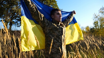 Military man in uniform stands with raised over head flag of Ukraine at countryside. Male ukrainian army soldier with lifted national banner in honor of victory against russian aggression. End of war