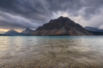 High-res landscape of Bow Lake, Alberta. Features dramatic storm clouds, turquoise glacial water, and rugged peak. Ideal for travel and outdoor branding.