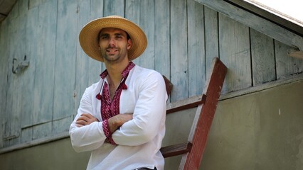 Portrait of smiling guy in straw hat against the background of his house. Young ukrainian man wearing an embroidered shirt looking into camera outdoor. Concept of national identity and patriotism