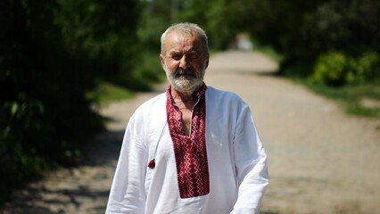 Elderly man wearing an embroidered shirt walking through rural road at sunny summer say. Senior male in traditional vyshyvanka going along country trail. Concept of national identity and patriotism