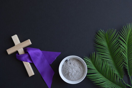 Wood cross with purple ribbon, bowl of ashes and palm leaves on a black background with copy space