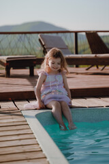 Cheerful girl dangling her legs in the pool. The little girl is very happy and joyful. Mermaid dress. A swim ring with a fish tail lies near the pool. Vertical photo.