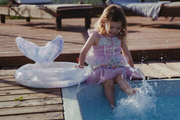 Cheerful girl dangling her legs in the pool. A little girl is sitting by the pool in a mermaid costume. A swim ring with a fish tail lies near the pool. Warm and sunny weather.