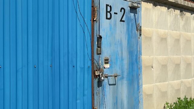 Blue metal door with padlock, weathered gate labeled B against concrete block wall, rusted hinges and chipped paint, overgrown weeds at perimeter of urban construction site security guard.