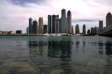 Obraz premium Wide panoramic view of Dubai Marina skyline across calm waterfront, modern skyscrapers reflecting daylight, showcasing architecture, coastal cityscape, travel destination.
