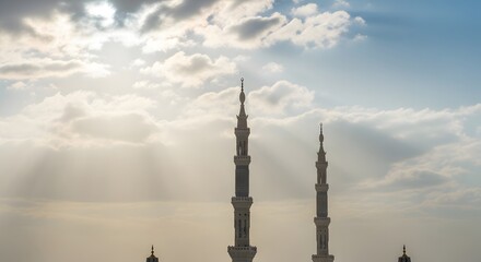 Obraz premium Sunlight filtering through clouds illuminates the towering minarets of a mosque, a symbol of Islamic architecture and spiritual devotion